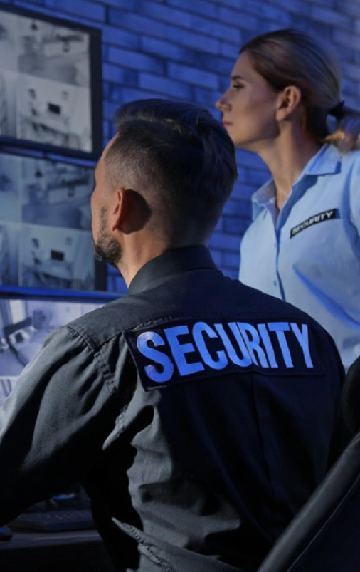Access Control Security Screening In a dark security control room, two guards, a man and a woman, attentively watch multiple CCTV surveillance monitors. The male guard is seated with his back to the camera, wearing a black uniform with