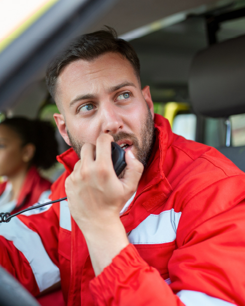 Alarm Response Service A focused male paramedic with a beard and short hair is sitting in the driver's seat of an ambulance. He is wearing a red and white jacket and is speaking into a two-way radio. In the blurred background, another person is visible in the passenger seat.