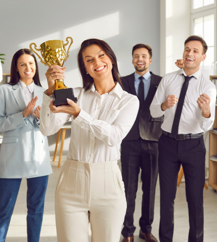 Award Winning Excellence A happy young businesswoman holds a large golden trophy while smiling and looking at the camera. She stands in an office with her team cheering behind her, with one woman on the left clapping and two men on the right celebrating enthusiastically. The group is celebrating an achievement or award.