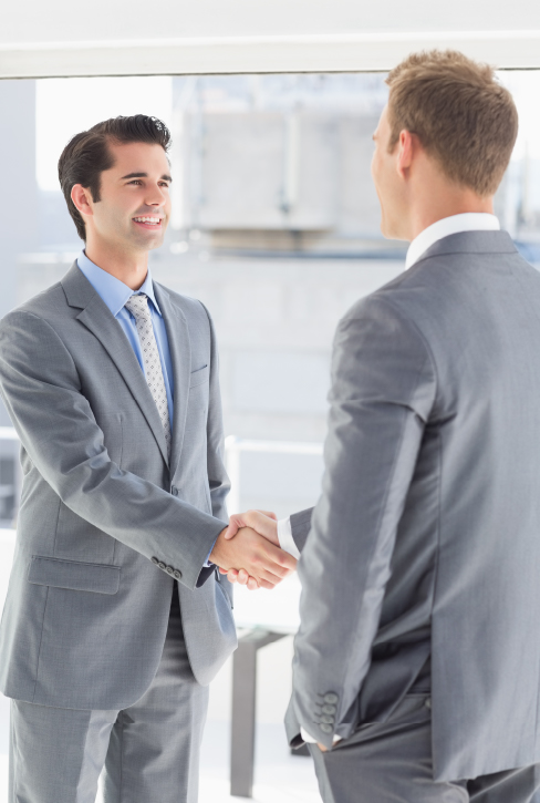 Government Approved Nationwide Two male businessmen in suits are shaking hands in front of a large window in an office building. The man on the left is smiling and facing the camera, while the man on the right has his back to the camera. The image suggests a business agreement or partnership.
