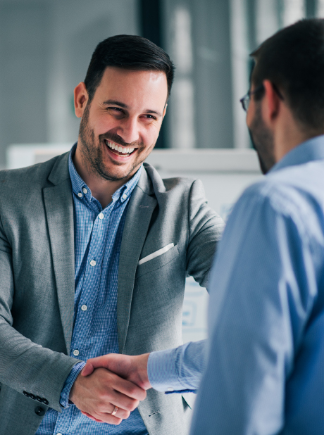 Local Expertise in Every City We Serve A man in a grey blazer smiles warmly as he shakes hands with another professional in a bright office setting. The focus is on the smiling man, conveying a sense of trust and a positive business agreement.