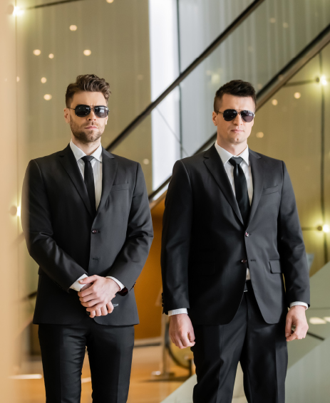 Corporate Hosting and VIP Security Two security personnel in black suits and sunglasses standing indoors, maintaining a professional and alert posture in a modern corporate setting.