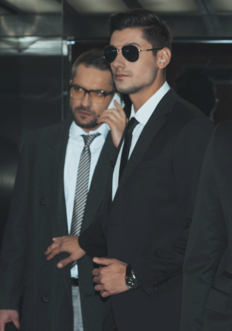 Rigorous Training and Quality Assurance Three professional security agents in black suits and sunglasses standing in an elevator, wearing earpieces and maintaining a serious, alert demeanor.