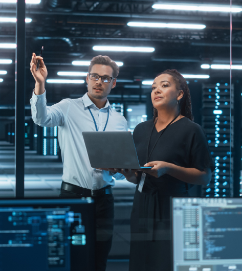 Security Risk Management Planning Two IT professionals discussing cybersecurity strategy in a data center, with one holding a laptop and computer monitors displaying code in the foreground.