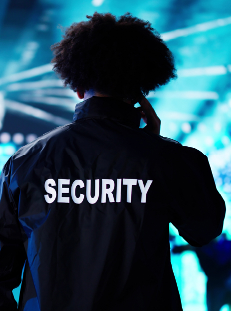 Security and Event Management A security guard wearing a black jacket labeled “SECURITY” on the back, standing in front of a brightly lit blue background while communicating through an earpiece.